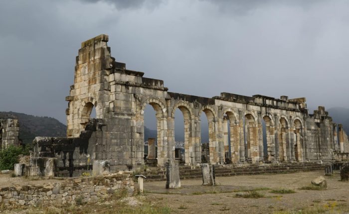 Ancient Roman ruins of Volubilis in Morocco, showcasing stone arches and dramatic cloudy sky.
