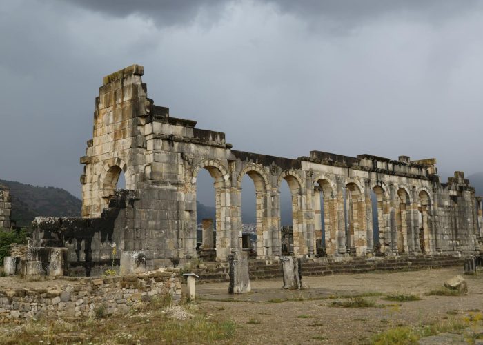 Ancient Roman ruins of Volubilis in Morocco, showcasing stone arches and dramatic cloudy sky.